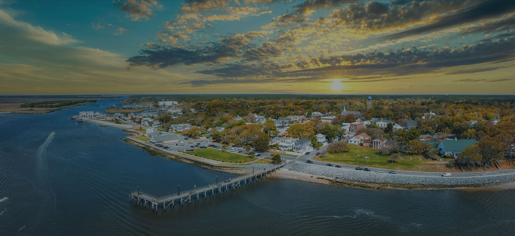 Aerial view of the Southport waterfront at sunset, with the pier reaching into the Cape Fear River and homes among autumn trees along the shore