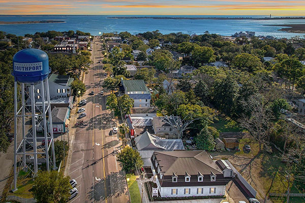 Aerial view of a downtown Southport street lined with trees and the blue Southport water tower, with the river beyond
