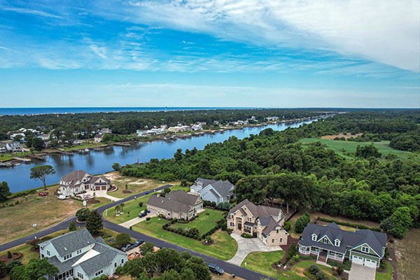 Aerial view of large single-family homes along a curving road beside a tidal creek, with the ocean on the horizon